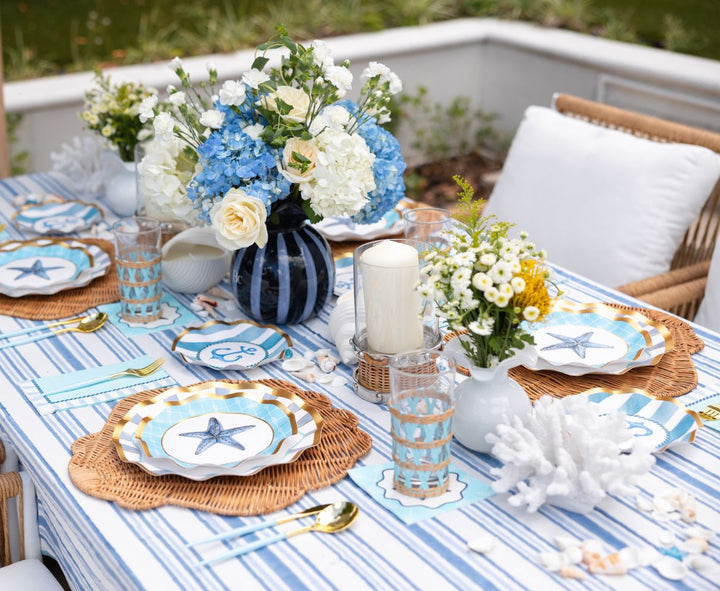 Create a stunning coastal-themed party with the Tidal Wave Table Setting by Sophistiplate—featuring blue and white striped linens, designer plates on wicker placemats, floral accents, candles, seashells, coral decor, and elegant glassware.