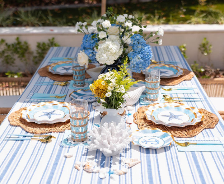 The Tidal Wave Table Setting by Sophistiplate creates a coastal vibe with blue and white striped tablecloth, starfish disposable plates, woven placemats, blue glassware, floral centerpieces, plus seashell and coral accents for outdoor dining.