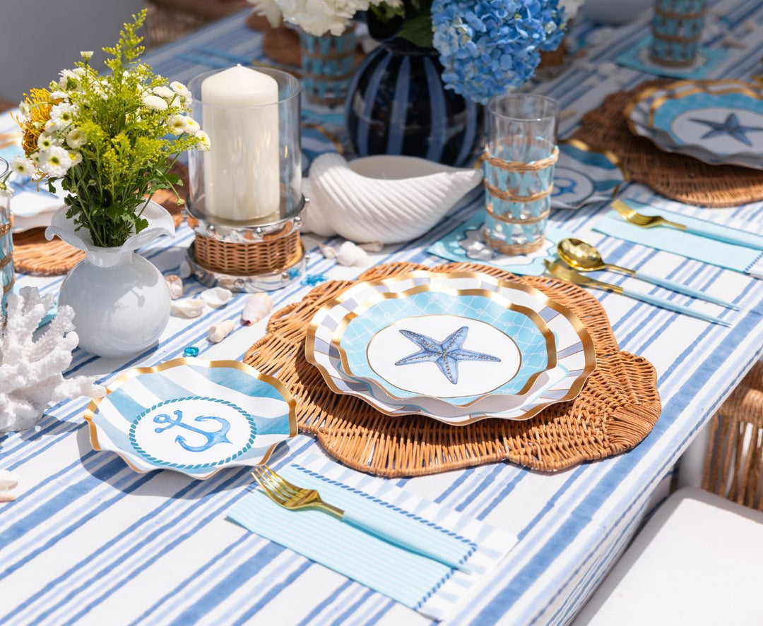 Create a seaside-inspired tablescape with the Sophistiplate Tidal Wave Table Setting—featuring blue and white stripes, wicker accents, starfish and anchor plates, gold cutlery, and a hydrangea centerpiece.