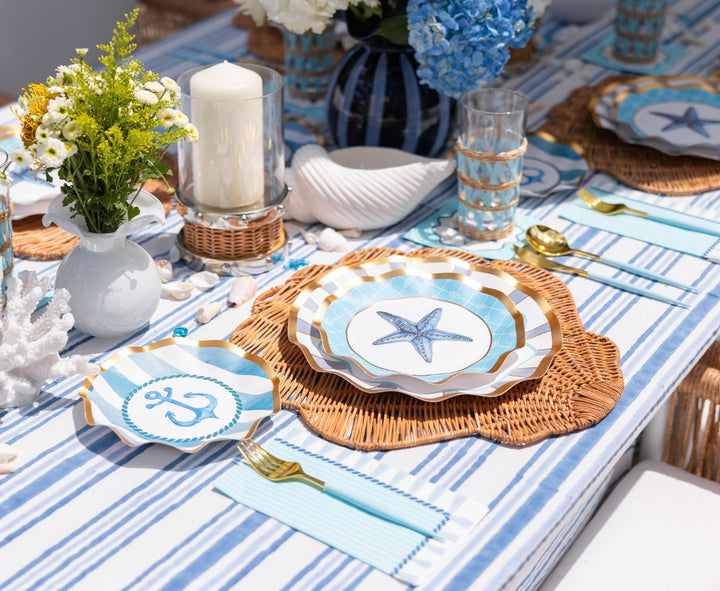 Create a seaside-inspired tablescape with the Sophistiplate Tidal Wave Table Setting—featuring blue and white stripes, wicker accents, starfish and anchor plates, gold cutlery, and a hydrangea centerpiece.