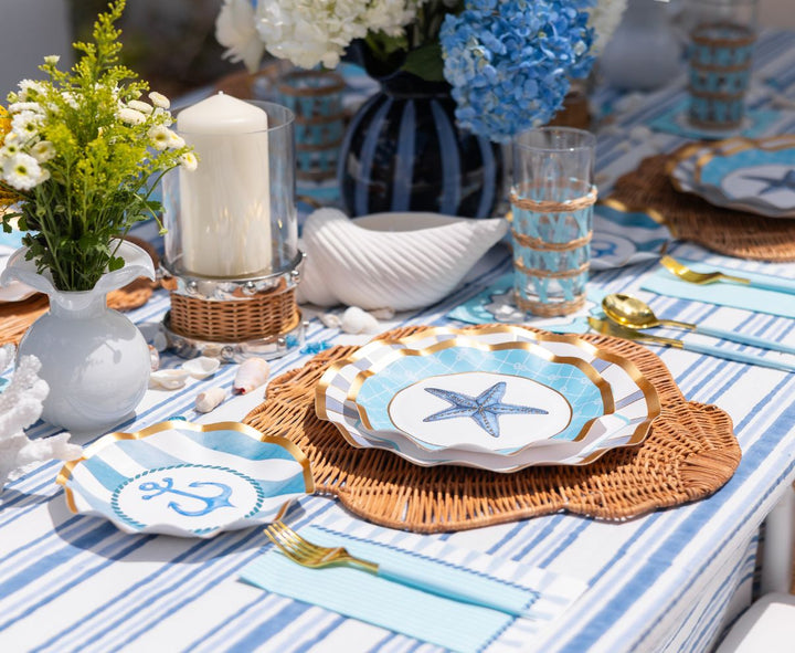 The Tidal Wave Table Setting by Sophistiplate features blue and white striped linens, starfish and anchor paper plates, gold utensils, wicker placemats, glassware, flowers, a white candle, and decorative seashells for a nautical tablescape.
