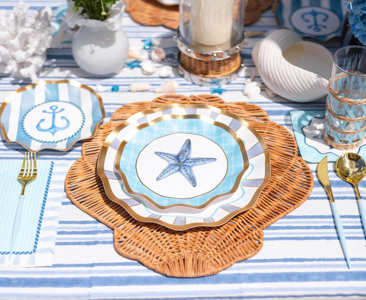 The Tidal Wave Table Setting by Sophistiplate creates a seaside vibe with blue and white plates accented in gold, wicker placemats, striped napkins, seashell decor, and gold cutlery for the perfect nautical-themed party tablescape.