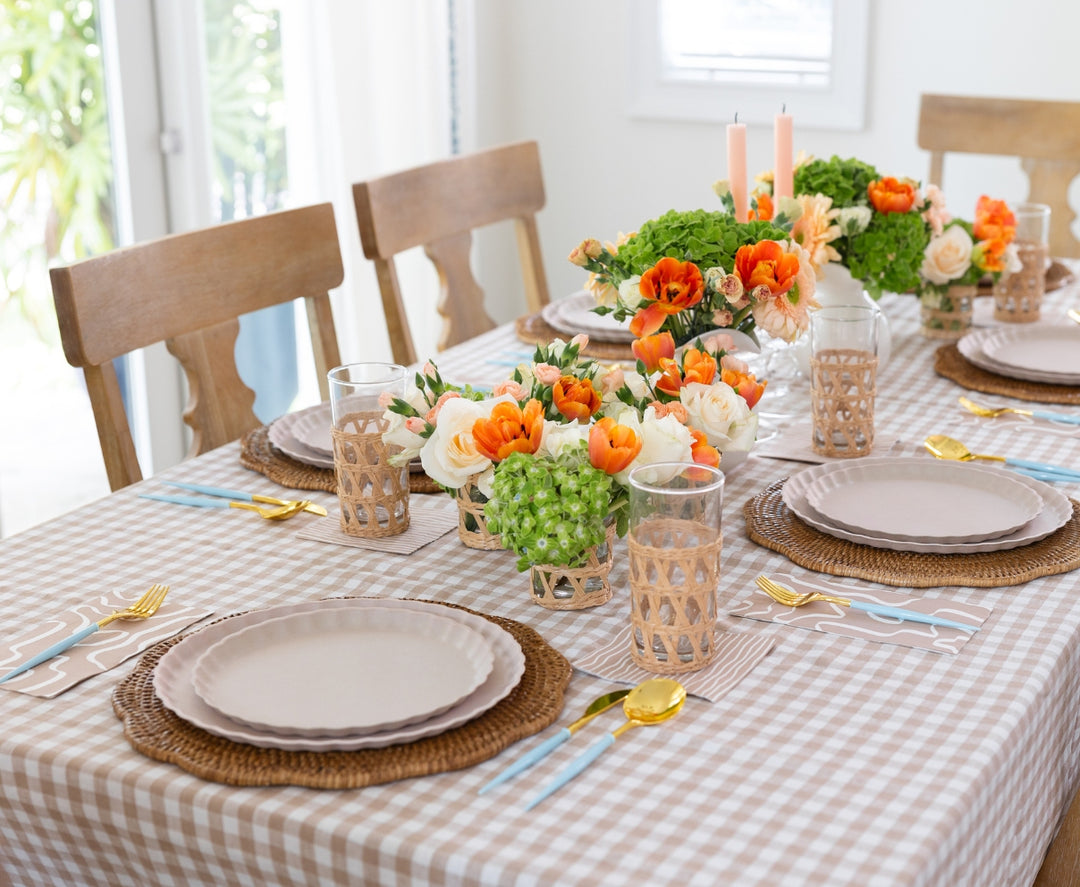A dining table set with beige plates, wicker placemats, glasses in wicker holders, and Sophistiplate Bella Cutlery Gold/Sky Blue Handle (24pkg), decorated with orange and white flowers and green foliage on a checkered tablecloth.