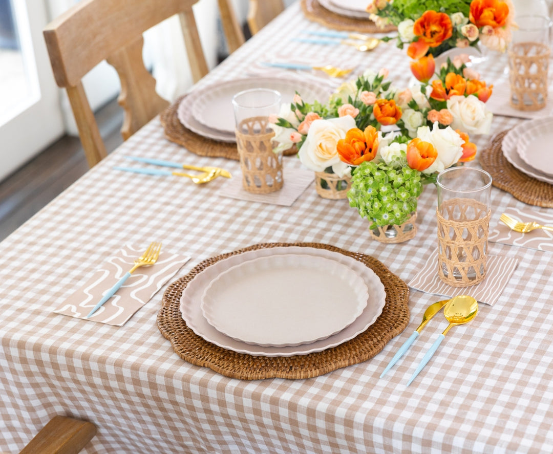 A dining table set with beige plates, gold cutlery, and rattan-holder glasses features Sophistiplate Guest Towel Eco Sand napkins, complementing a checkered tablecloth and orange, white, and green floral arrangements.