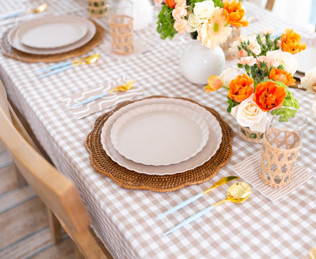 A table is set with a checkered tablecloth, woven placemats, cream plates, gold utensils, glasses, and Sophistiplate's Cocktail Napkin Eco Sand/20 pkg paper napkins for convenience, plus vases of orange and white flowers as centerpieces.