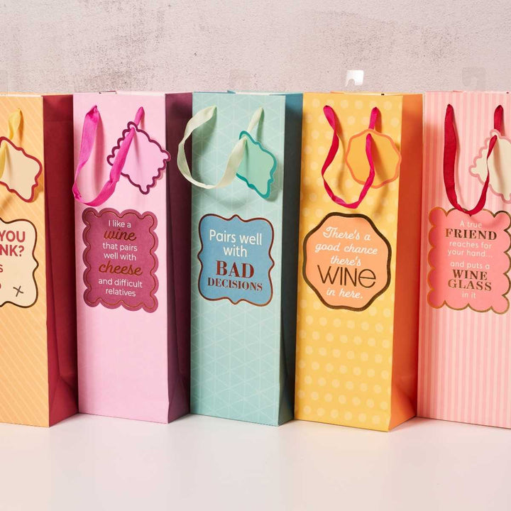 Five Drinks On Me Good Chance Wine Bags with humorous wine, cheese, and friendship phrases feature decorative tags and ribbon handles. These colorful, reusable gift bags are lined up against a light wall.
