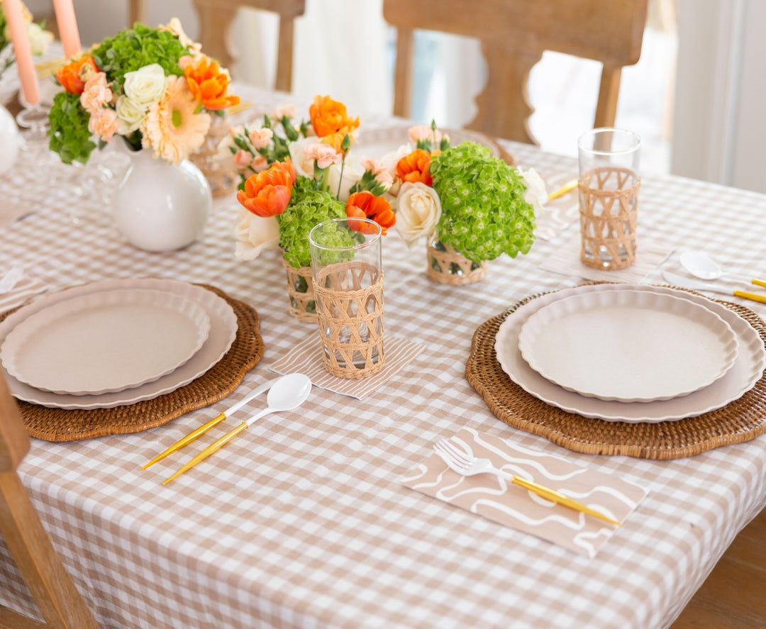 A dining set features a checkered beige tablecloth, woven placemats, beige plates, gold cutlery, Sophistiplate Guest Towel Eco Sand napkins, glasses with wicker holders, and vases of orange, cream, and green flowers as centerpieces.