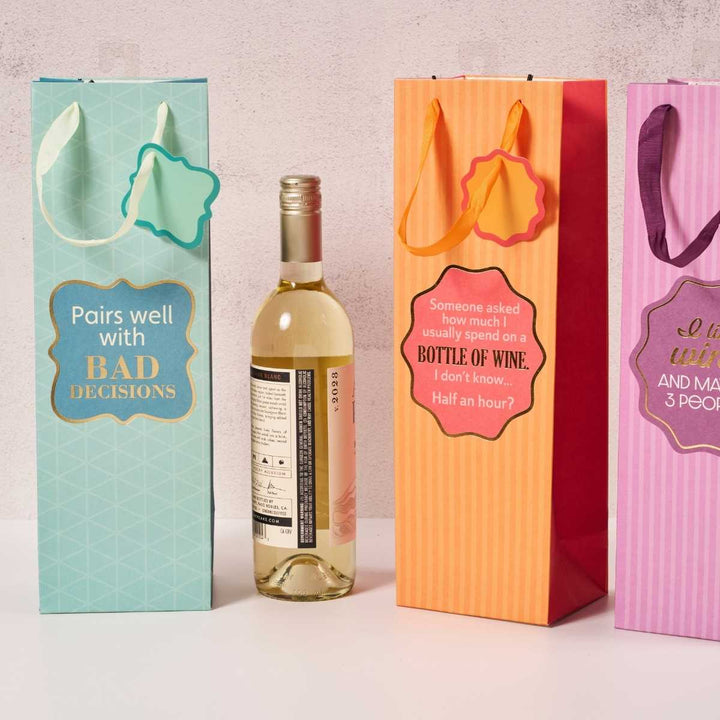 The Drinks On Me "Bad Decisions" Wine Bag features a colorful, funny design with the label "Pairs well with bad decisions," making it a unique reusable wine bag or hostess gift. Shown next to a bottle of white wine on a light textured background.