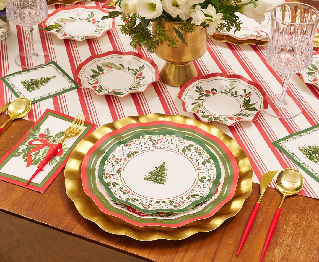 Create a festive table setting with gold-rimmed plates adorned with Christmas tree designs, gold cutlery featuring red handles, crystal glasses, and holiday-themed cards arranged atop a red and white striped tablecloth. Sophistiplate’s “Home for the Holidays” Guest Towels (20pk) serve as elegant napkins at each place setting, while flowers displayed in a gold vase complete the holiday look.