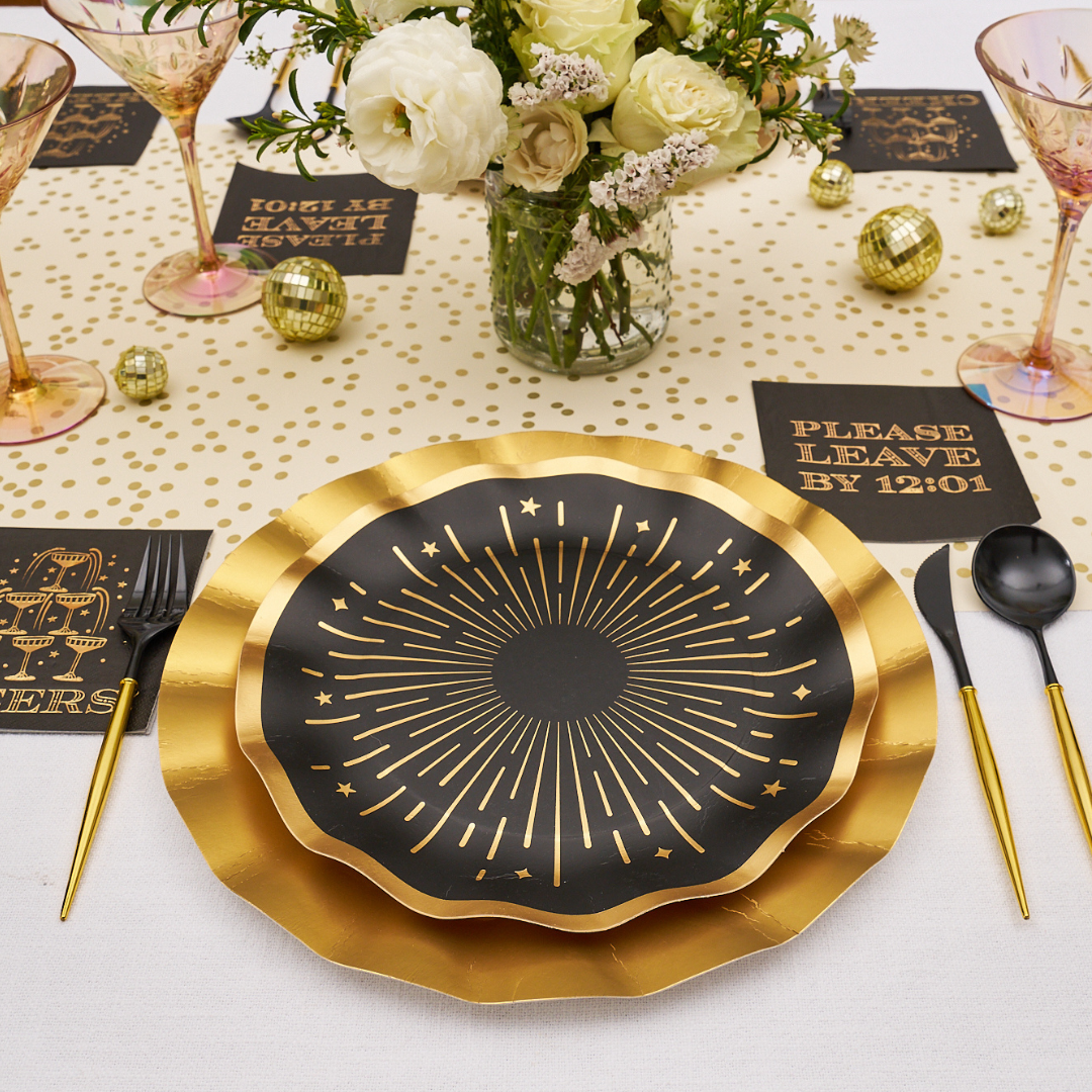 Midnight Sparkle Table Setting featuring a layered place setting with a black, starburst-patterned plate on a gold charger, black napkins reading PLEASE LEAVE BY 12:01 and CHEERS, black and gold cutlery, and small gold disco ball decorations.
