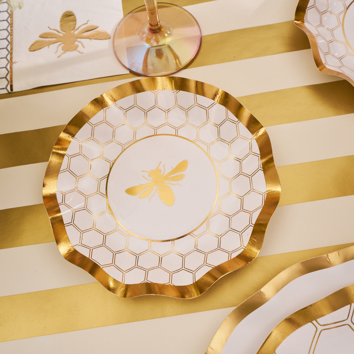 A Sophistiplate Honeybee White Wavy Paper Appetizer/Dessert Bowl from the 8-pack sits on a white and gold striped table, with matching elegant plates, napkin, and glass partially visible nearby.