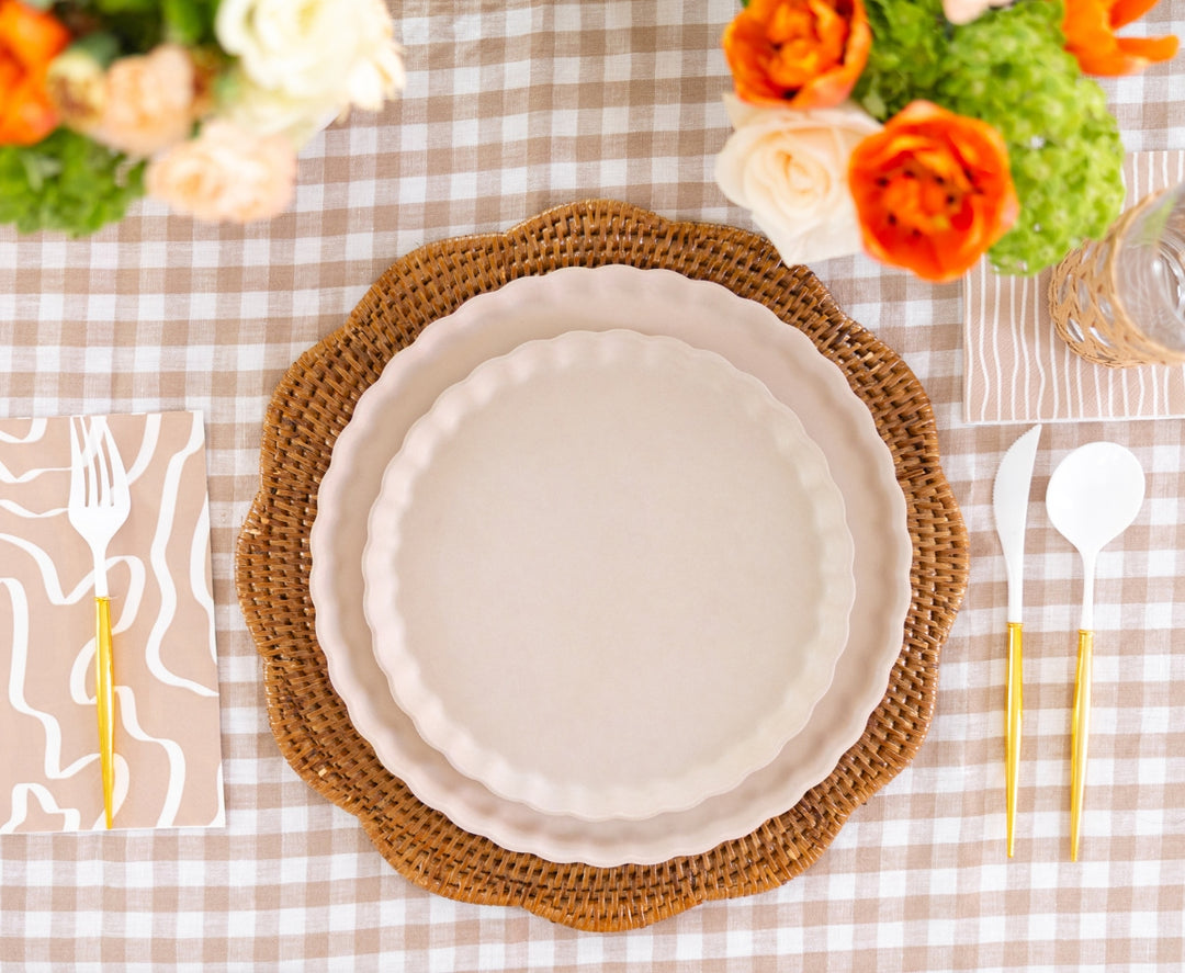 A table setting features two stacked white scalloped plates on a woven charger, gold utensils, Sophistiplate Guest Towel Eco Sand napkins, and vases of orange, white, and green flowers on a beige gingham tablecloth.