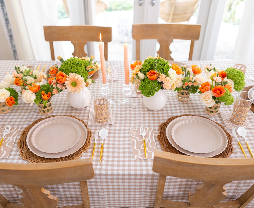 A dining table set for two with a tan checkered tablecloth, white plates on wicker chargers, gold cutlery, Sophistiplate Guest Towel Eco Sand napkins, two lit candles, and flower arrangements in vases and woven holders.