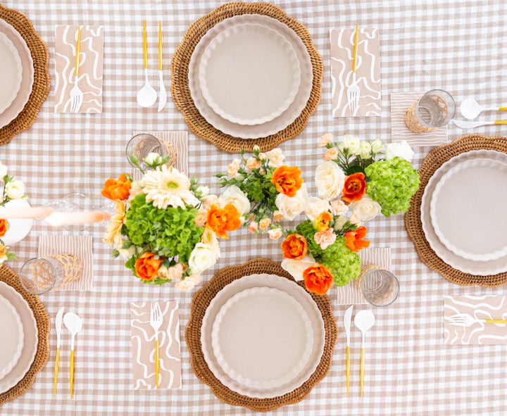 A table for six features beige plates on woven placemats, gold and white cutlery, glasses, vibrant floral centerpieces, and Sophistiplate’s Guest Towel Eco Sand paper napkins on a beige gingham tablecloth.