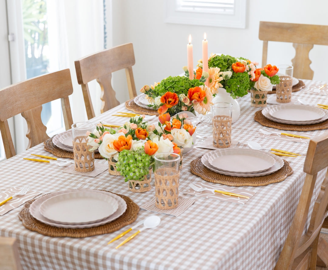 The Sophistiplate Eco-Friendly Table Setting - Sand features compostable plates, woven placemats, gold and white cutlery, glasses in woven holders, floral centerpieces in orange, white, green hues, and two lit candles for a table set for four.