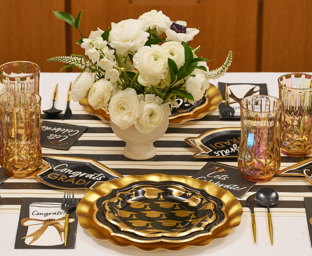 The Classic Grad Table Setting by Sophistiplate includes designer gold and black disposable plates, gold utensils, rose glasses, “Congrats Grad!” signs, a white floral centerpiece, and a striped table runner for a festive celebration.