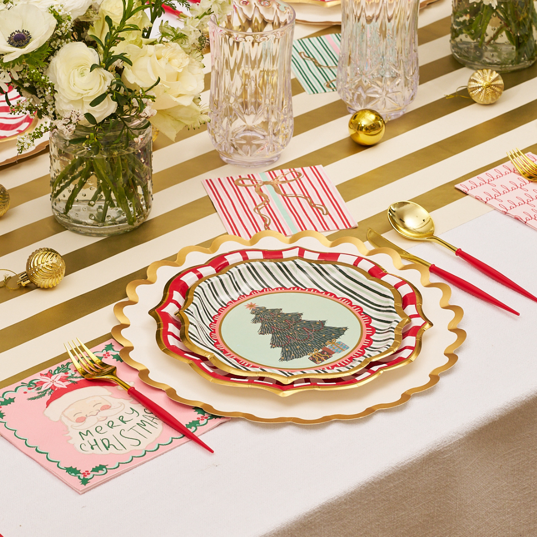 Celebrate in style with a festive table setting featuring a gold-striped cloth, a floral centerpiece, and Sophistiplate Tis the Season Salad Plates (8pk), adorned with a charming Christmas tree design. The arrangement is completed with gold cutlery accented by red handles, crystal glasses, decorative ornaments, cheerful holiday napkins, and festive paper plates—perfect for serving appetizers or sides.