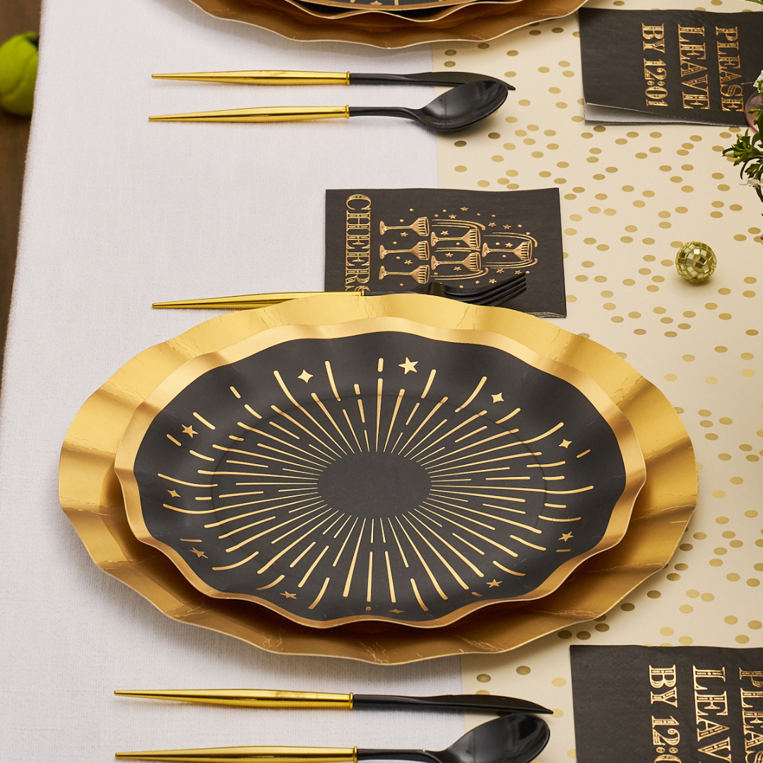 Midnight Sparkle Table Setting featuring black and gold scalloped disposable plates with a starburst design, matching napkins with CHEERS and champagne glass illustrations, and black plastic cutlery with gold handles on a table.