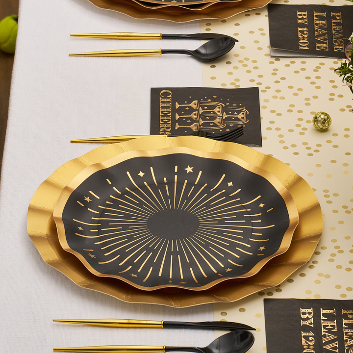 Midnight Sparkle Table Setting featuring black and gold scalloped disposable plates with a starburst design, matching napkins with CHEERS and champagne glass illustrations, and black plastic cutlery with gold handles on a table.