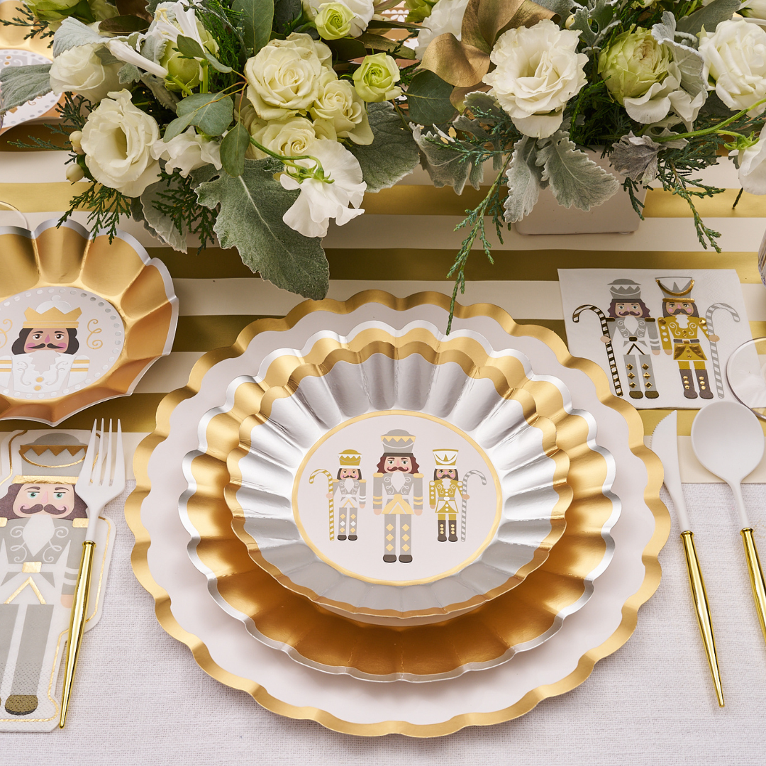 The Gilded Nutcracker Table Setting by Sophistiplate showcases gold and white scalloped plates with nutcracker designs, white cutlery, and floral arrangements atop a striped gold runner for a festive holiday table.