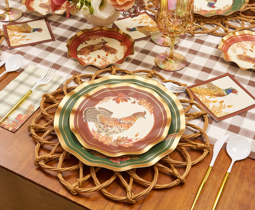 A fall-themed table setting featuring Sophistiplate Woodland Harvest Salad Plates (8pk) with charming bird prints, accompanied by woven placemats, plastic cutlery, napkins, and glasses arranged on a brown plaid tablecloth. A vase of flowers completes the inviting Thanksgiving-inspired atmosphere.