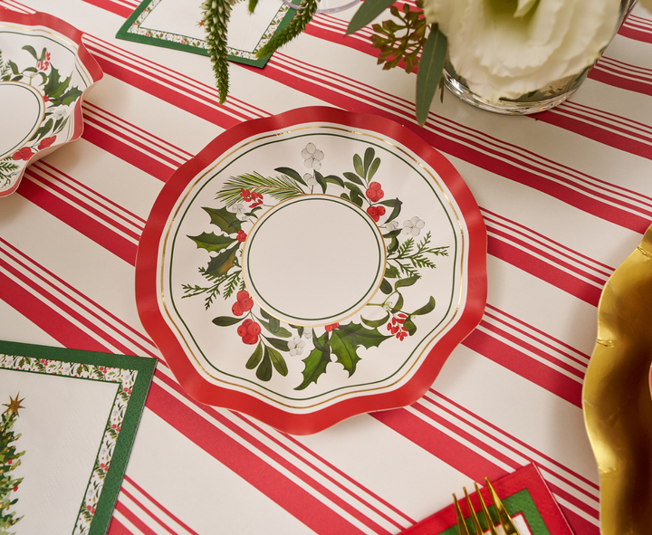 A decorative plate adorned with holly and berries rests on a red and white striped tablecloth, surrounded by festive place settings featuring napkins, a gold charger, Sophistiplate’s Home for the Holidays Appetizer/Dessert Bowls (8pk) with wavy edges, and a holiday floral arrangement.
