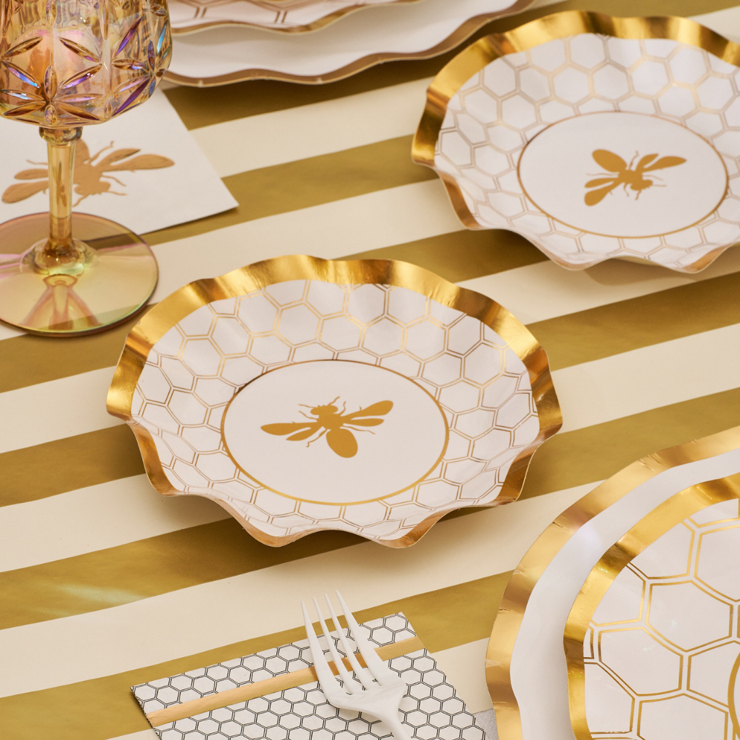 A table setting featuring Sophistiplate’s Honeybee White Wavy Paper Appetizer/Dessert Bowls, a matching napkin, gold-striped tablecloth, pink glass, and a white plastic fork.