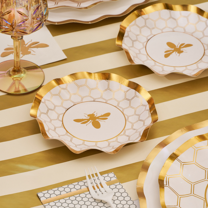 A table setting featuring Sophistiplate’s Honeybee White Wavy Paper Appetizer/Dessert Bowls, a matching napkin, gold-striped tablecloth, pink glass, and a white plastic fork.