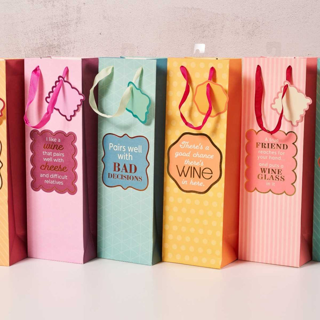 Five vibrant "Bad Decisions" wine bags by Drinks On Me stand in a row—pink, blue, yellow, orange, and pink striped—each featuring playful wine sayings and tags. A funny hostess gift or unique reusable bag for wine lovers.