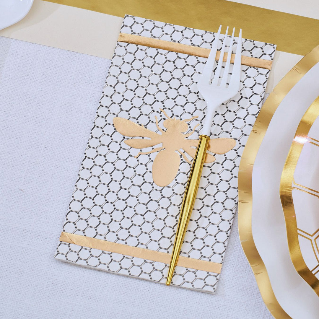 The White Honeybee Table Setting by Sophistiplate features a napkin adorned with a honeycomb pattern and gold bee motif, elegantly holding a white fork and gold spoon, arranged next to sophisticated party tableware including white disposable plates with gold rims on a refined white and gold tablescape.