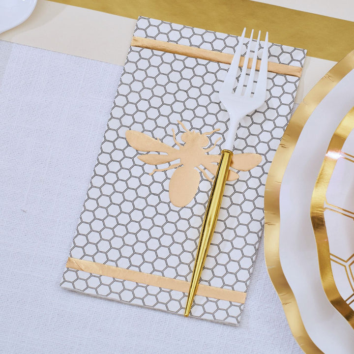 The White Honeybee Table Setting by Sophistiplate features a napkin adorned with a honeycomb pattern and gold bee motif, elegantly holding a white fork and gold spoon, arranged next to sophisticated party tableware including white disposable plates with gold rims on a refined white and gold tablescape.