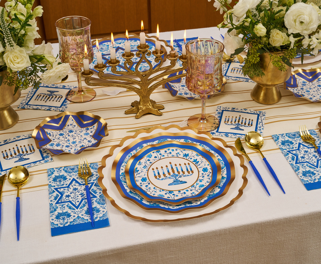 The Sophistiplate Hanukkah Toile Table Setting features elegant blue, white, and gold designer paper plates, menorah-themed napkins, gold cutlery, floral centerpieces, and a lit menorah centerpiece—all combining to create a festive tablescape perfect for your holiday celebration.