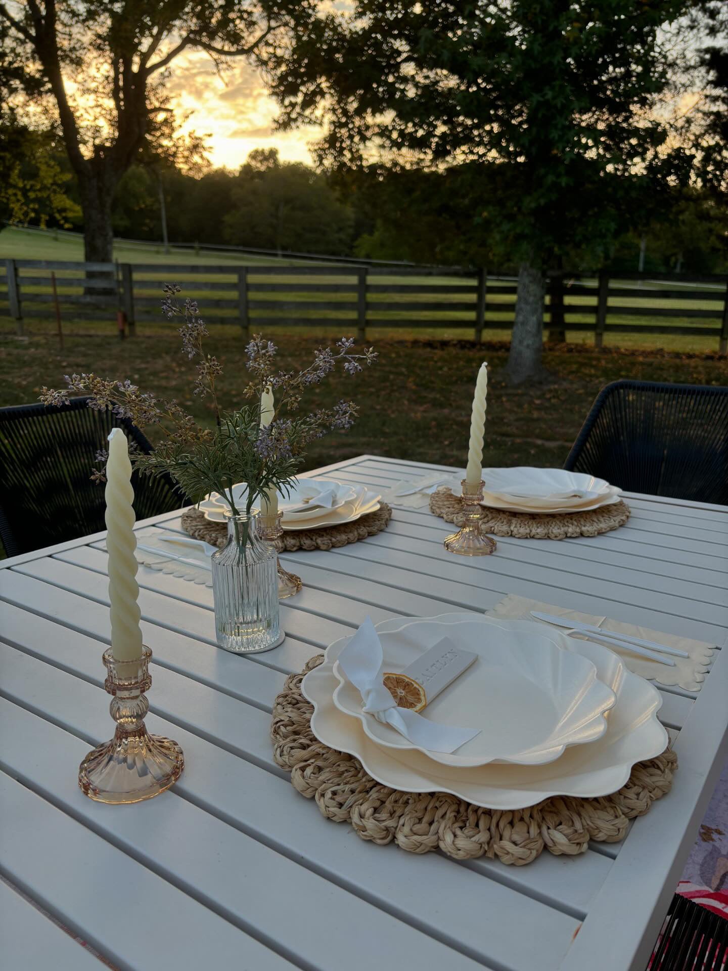 The Sophistiplate Eco-Friendly Table Setting- Cream features tableware for four, gold cutlery, spiral candles in glass holders, and a wildflower vase on an outdoor table set at sunset near trees and a wooden fence.