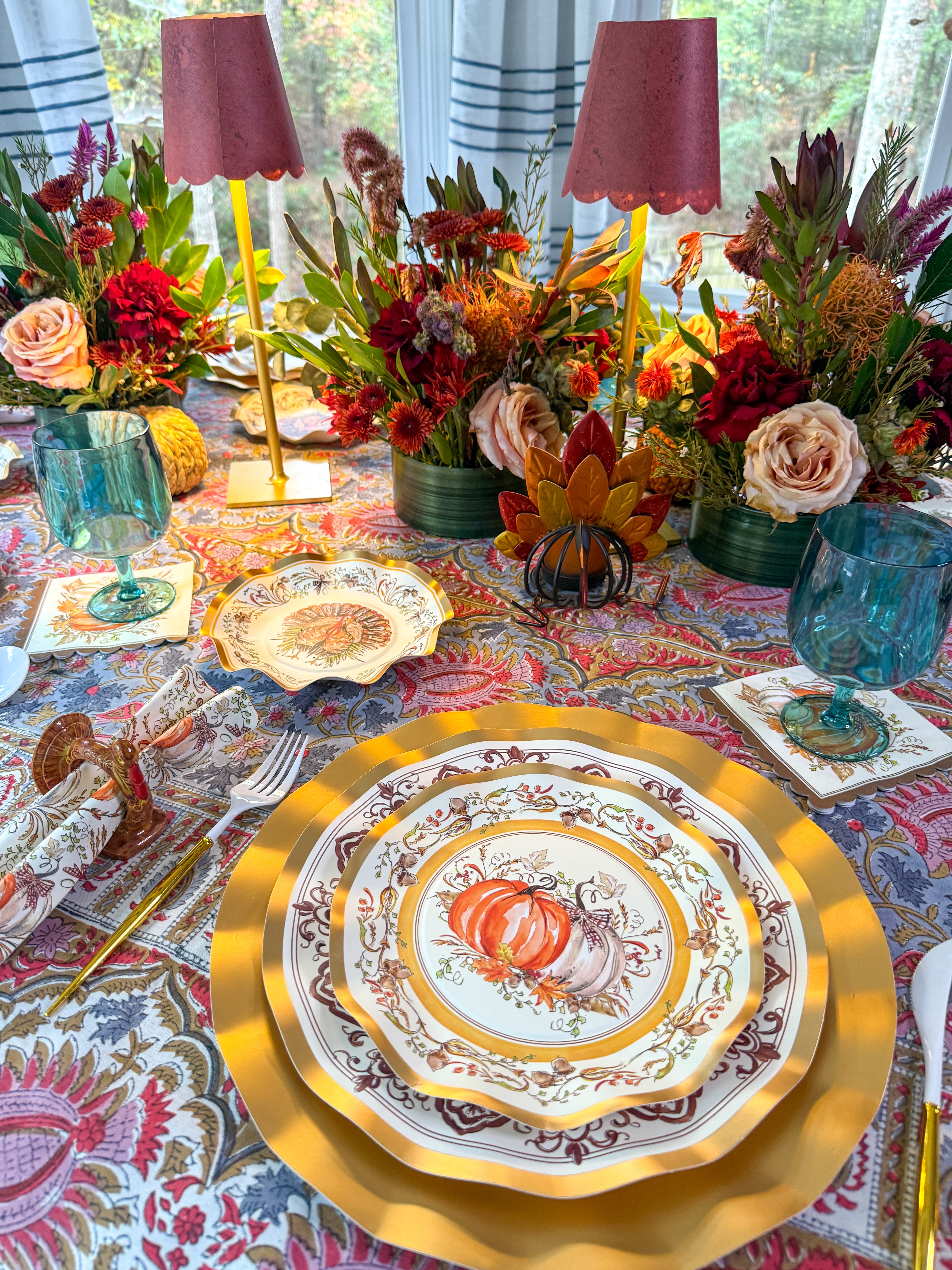 Turkey & Gourds: Thanksgiving Table for 16 set on a patterned tablecloth with gold chargers, teal glasses, and floral centerpieces with candles, featuring paper plates decorated with pumpkin illustrations.