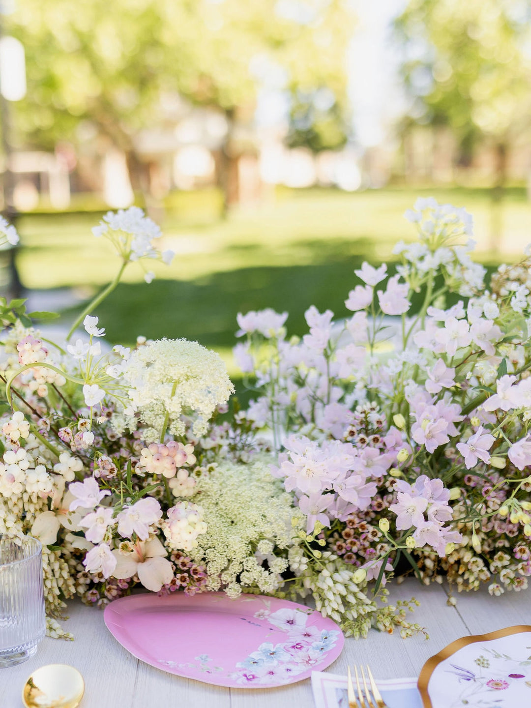 An outdoor table is set with a Sophistiplate Egg Salad Plate Charming Easter Assorted (8-pack), accompanied by a glass, utensils, and a lush centerpiece of white and light pink flowers. In the background, a vibrant green park scene appears softly out of focus.