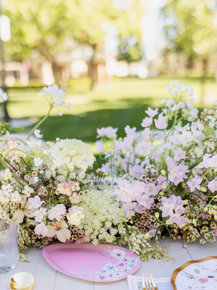 An outdoor table is set with a Sophistiplate Egg Salad Plate Charming Easter Assorted (8-pack), accompanied by a glass, utensils, and a lush centerpiece of white and light pink flowers. In the background, a vibrant green park scene appears softly out of focus.
