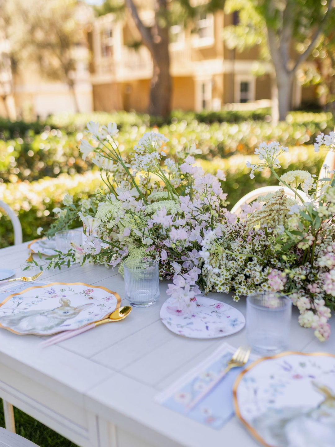 The Charming Easter Table Setting by Sophistiplate features a white outdoor table adorned with designer disposable plates showcasing elegant floral patterns, complemented by gold-rimmed cutlery, clear glasses, and a lush centerpiece of white and pale purple flowers—all beautifully arranged amidst greenery and sunlight.