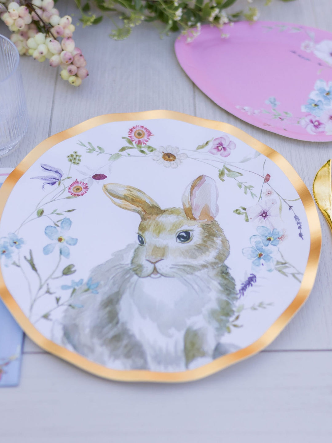 Introducing the Wavy Dinner Plate Charming Easter - 8pkg by Sophistiplate—a beautifully designed plate adorned with a watercolor rabbit nestled among vibrant flowers. Set atop a light wooden table with a gold fork and coordinating pink floral plate, this delightful piece captures the whimsical spirit of Easter. The elegant wavy edge and delicate gold foil trim add an extra touch of sophistication, making it perfect for festive gatherings.