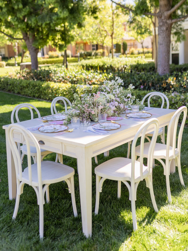 An elegant outdoor table set for six, featuring white chairs and Sophistiplate’s Wavy Dinner Plate Charming Easter (8-pack) with gold foil trim, paired with glasses and a lush floral centerpiece. The table is arranged on green grass, surrounded by trees and shrubs under bright sunlight.