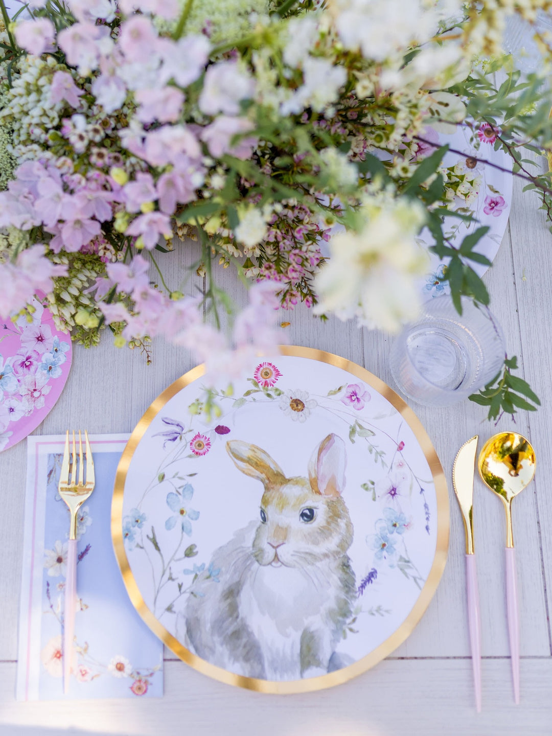 The Charming Easter Table Setting by Sophistiplate features designer disposable plates adorned with a rabbit illustration, gold-rimmed cutlery, a floral napkin, a clear glass, and a bouquet of pink and white flowers arranged on a light wooden table.