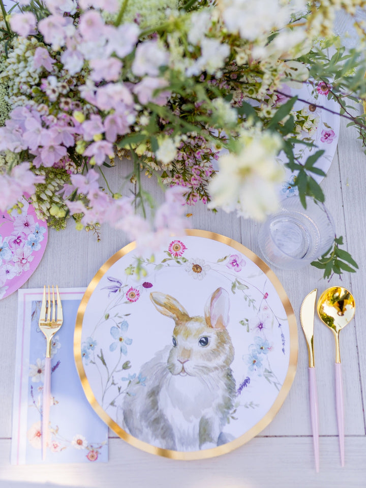 The Charming Easter Table Setting by Sophistiplate features designer disposable plates adorned with a rabbit illustration, gold-rimmed cutlery, a floral napkin, a clear glass, and a bouquet of pink and white flowers arranged on a light wooden table.