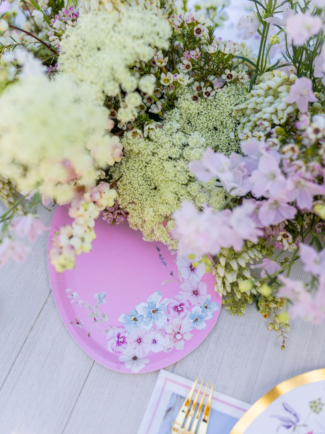 A “Charming Easter Table Setting” by Sophistiplate takes center stage on a white table, its pink designer disposable plate adorned with floral designs and surrounded by lush white and light pink blooms. A gold fork and knife rest at the bottom right corner, adding an elegant touch to this enchanting party-in-a-box arrangement.