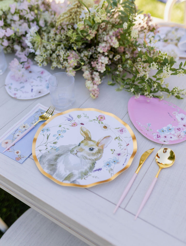 A Charming Easter Table Setting by Sophistiplate features designer disposable plates with floral patterns and a bunny illustration, complemented by pink placemats, gold and pink cutlery, and finished with a bouquet of flowers as the centerpiece.