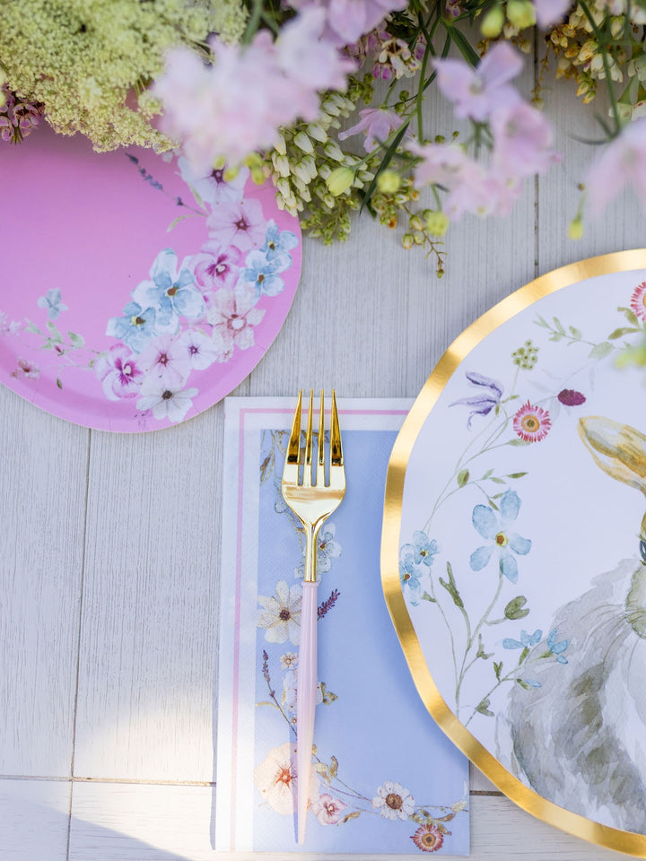 A close-up of the Charming Easter Table Setting by Sophistiplate, showcasing designer disposable plates with floral patterns, a gold fork resting on a pastel blue napkin, and pink flowers artfully arranged along the top edge.