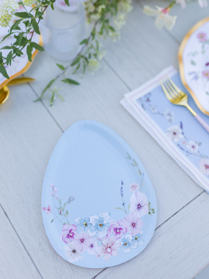 A set of Sophistiplate’s Wavy Dinner Plate Charming Easter plates, featuring pastel blue, egg-shaped designs with delicate florals and a subtle gold foil trim, is elegantly arranged on a light wooden spring table alongside gold utensils, coordinating floral plates, and touches of greenery in the background.