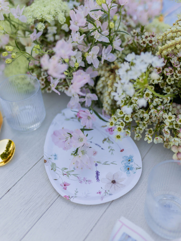 The Charming Easter Table Setting by Sophistiplate is beautifully displayed on a white wooden table, featuring designer disposable plates with floral patterns, accented by pastel flowers, clear glasses, and a gold spoon. Bathed in soft natural light, the elegant springtime scene is perfect for hosting an effortless party-in-a-box celebration.