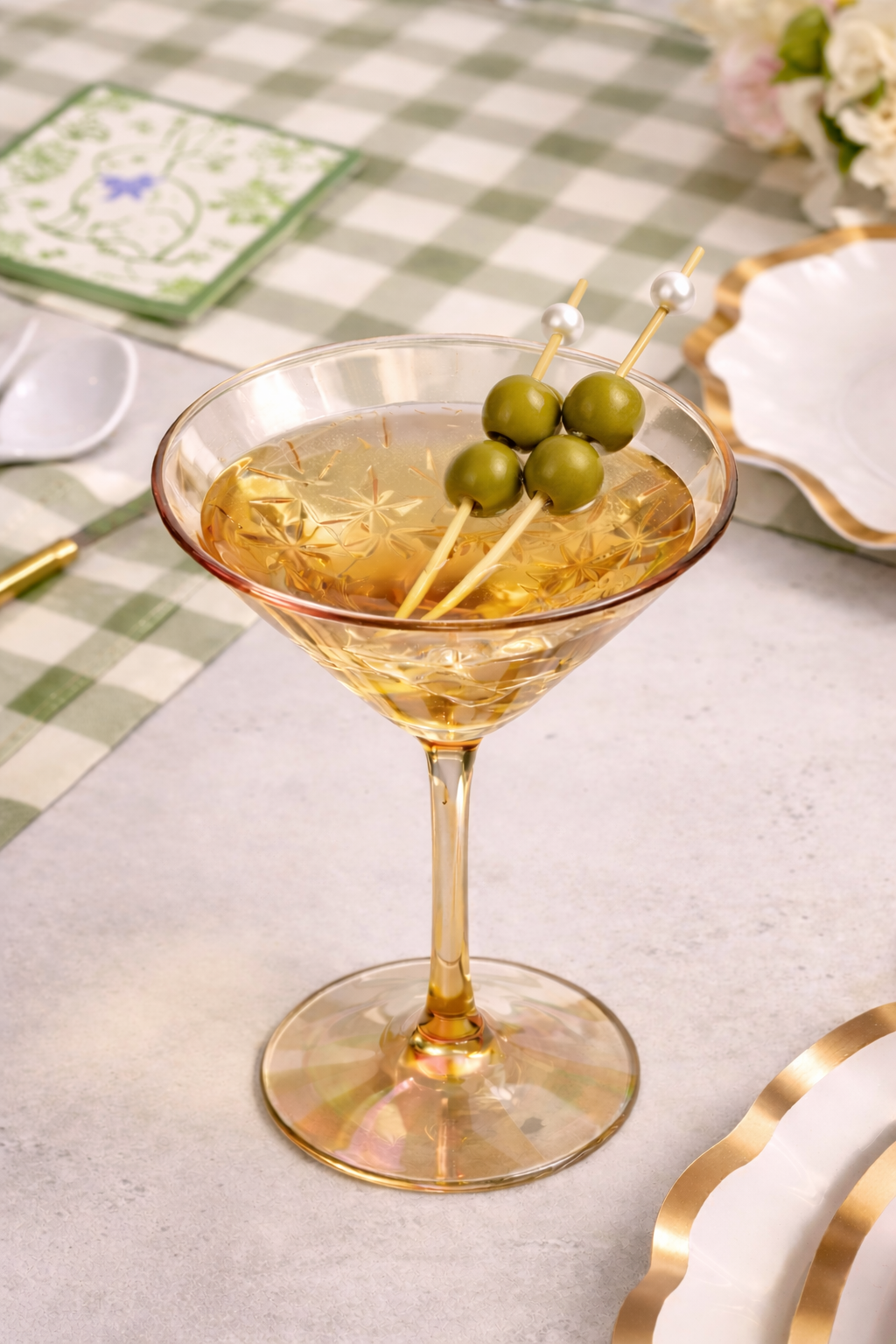 A martini glass with a light golden cocktail and three green olives sits on a green checkered tablecloth with Sophistiplate LLC’s Ultimate Easter Tablescape for 16: Easter Toile, creating an elegant setting.