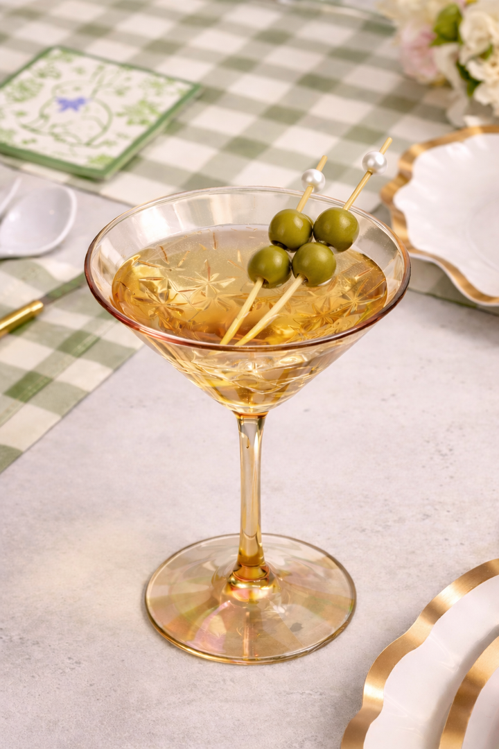 A martini glass with a light golden cocktail and three green olives sits on a green checkered tablecloth with Sophistiplate LLC’s Ultimate Easter Tablescape for 16: Easter Toile, creating an elegant setting.