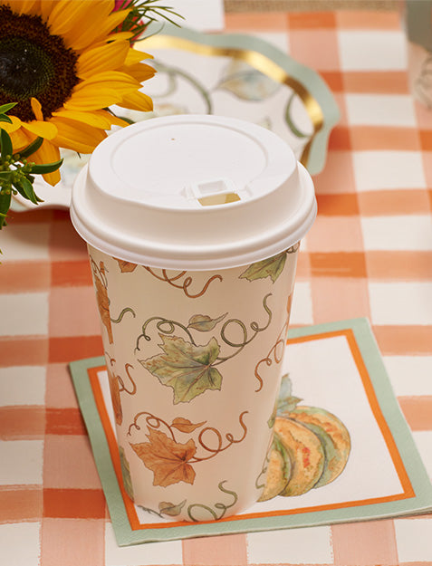 A paper coffee cup with autumn leaf designs sits on Sophistiplate’s Bright & Thankful Cocktail Napkins (26pk), atop an orange plaid tablecloth near a sunflower arrangement, creating the perfect setting for an autumn gathering.