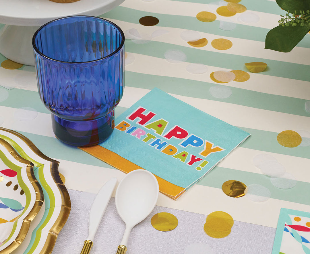 A blue glass, white plastic utensils, and Sophistiplate ‘Happy Birthday’ Cocktail Napkins (26pk) on a striped tablecloth with gold and white confetti scattered around create the perfect setting for your birthday decorations.